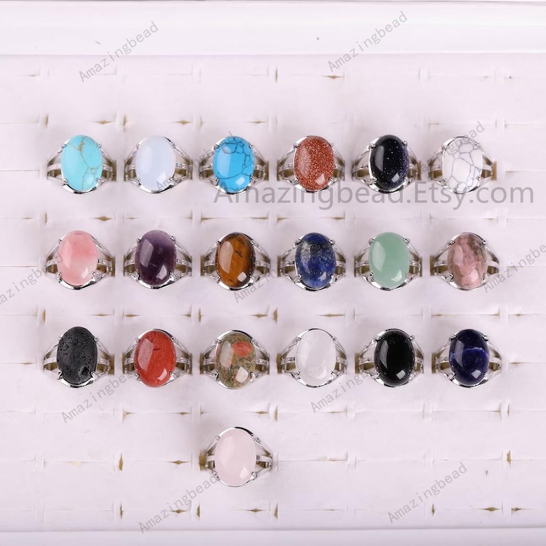 May include: A display of 18 oval-shaped gemstone rings with silver settings. The rings are arranged in a grid pattern on a white surface. The gemstones are various colors, including pink, red, green, blue, black, white, and brown.