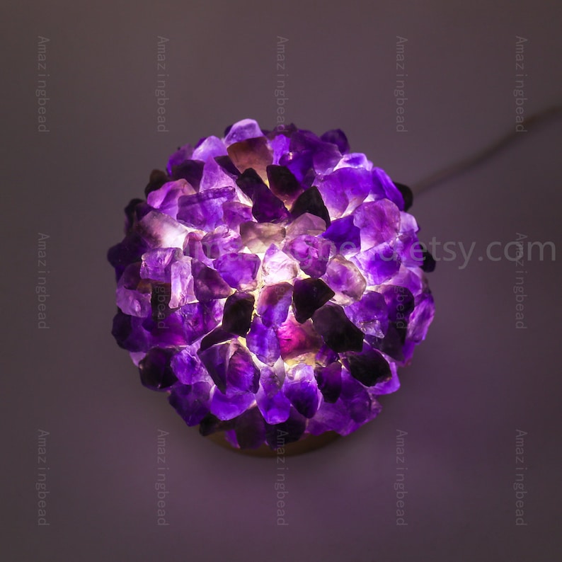 K&ouml;nnte beinhalten: Eine violette Amethyst-Kristalllampe mit einem warmwei&szlig;en Licht, das durch die Kristalle scheint.