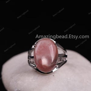 May include: A silver ring with a large oval-shaped pink gemstone. The ring has a delicate design with three bands that wrap around the stone.