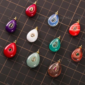 Puede incluir: Una colección de colgantes en forma de lágrima en varios colores, incluyendo rojo, morado, blanco y verde. Cada colgante presenta una argolla dorada y una gema central o elemento decorativo. Los colgantes se muestran sobre una superficie de cuadrícula negra.