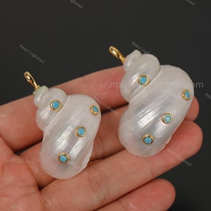 May include: Two white, pearlescent spiral shell pendants featuring small, round, bright blue stones set in gold-toned metal bezels. Each shell has a gold-toned metal loop at the top for attachment, showcasing a smooth, glossy, and iridescent surface.