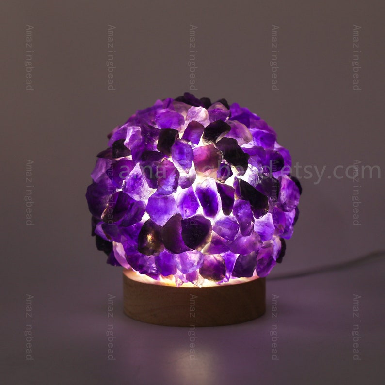 K&ouml;nnte beinhalten: Eine violette Amethyst-Kristalllampe mit einem Holzsockel. Die Lampe ist eingeschaltet und die Kristalle leuchten.
