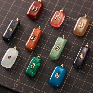 May include: A collection of rectangular gemstone pendants in various colors, including red, green, black, and blue. Each pendant has a gold-colored bail and a contrasting gemstone inlay. The pendants are arranged on a dark surface.