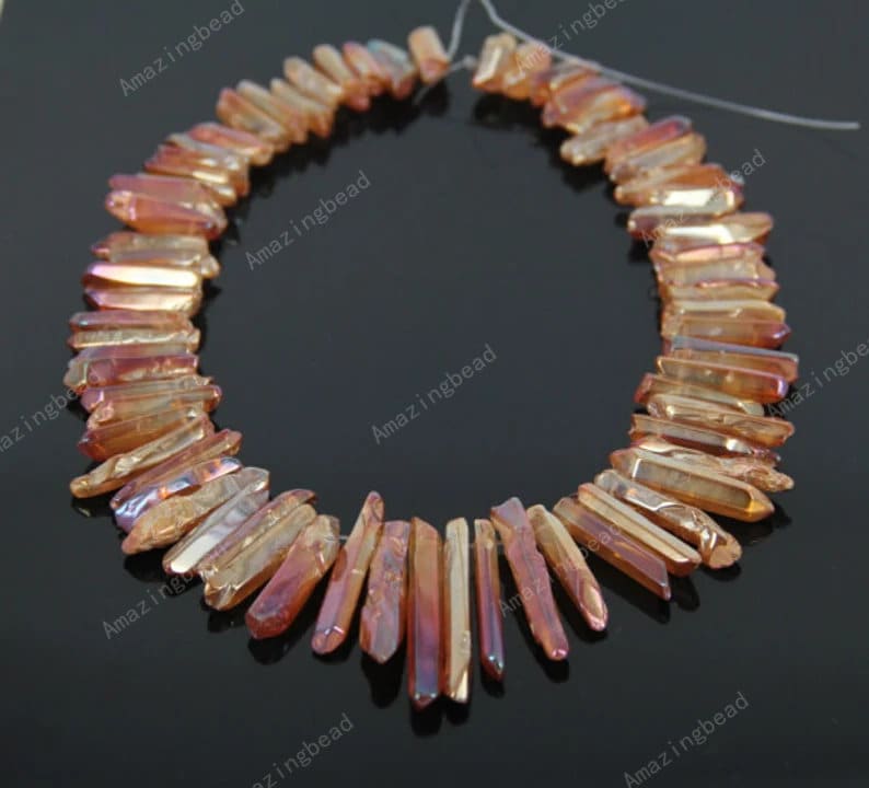 May include: A strand of orange and gold iridescent crystal point beads. The beads are faceted and have a pointed shape. The beads are strung on a clear thread.
