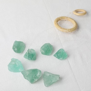May include: A crystal wind chime with seven light green fluorite crystals hanging from a wooden ring. The crystals are suspended from a clear string.