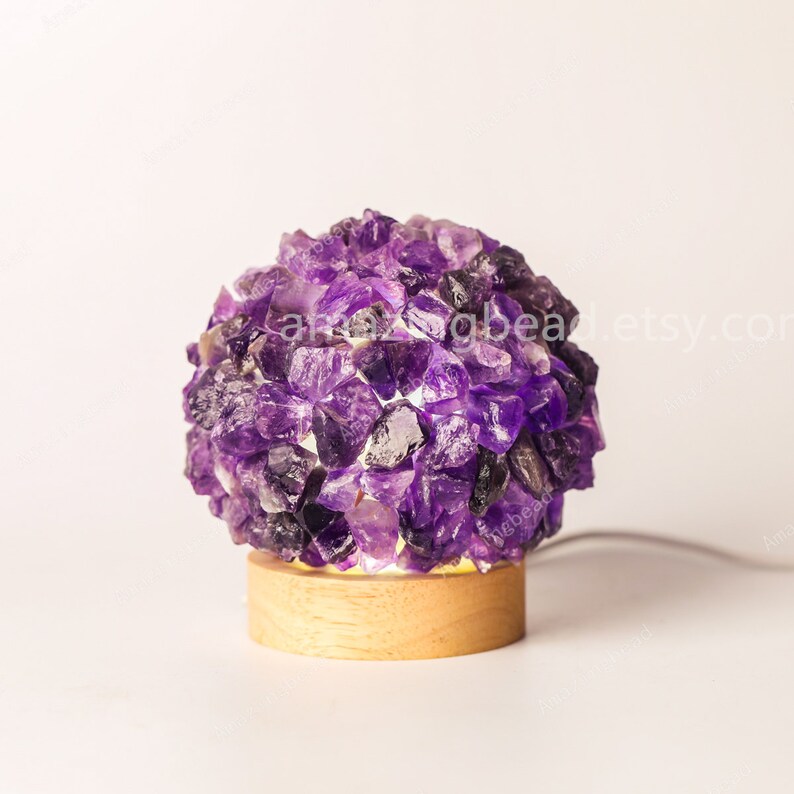 K&ouml;nnte beinhalten: Eine violette Amethyst-Kristalllampe mit einem Holzsockel. Die Lampe ist eingeschaltet und die Kristalle leuchten.