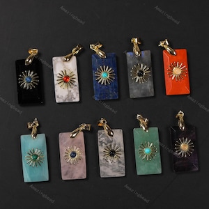 May include: A collection of rectangular gemstone pendants in various colors, including black, white, blue, gray, and red. Each pendant features a gold-colored sunburst design and a gold-colored bail for hanging.