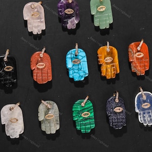 May include: A collection of gemstone pendants in the shape of the Hamsa hand. The pendants are in various colors, including clear, purple, green, red, blue, and orange. Each pendant features a gold-colored bail and an eye-shaped accent.