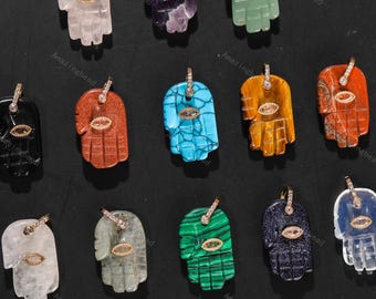 May include: A collection of gemstone pendants in the shape of the Hamsa hand. The pendants are in various colors, including clear, purple, green, red, blue, and orange. Each pendant features a gold-colored bail and an eye-shaped accent.