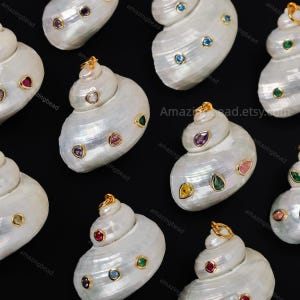 Peut inclure: Une collection de pendentifs en coquillage blanc irisé, chacun doté d'une bélière en métal doré sur le dessus et de plusieurs petites pierres précieuses facettées et colorées incrustées dans la surface du coquillage en diverses formes.