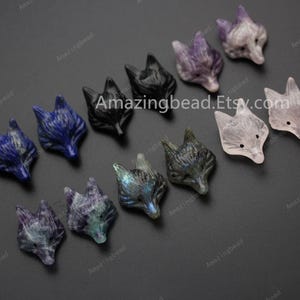 Könnte beinhalten: Eine Sammlung von 10 geschnitzten Fuchs-Kopf-Anhängern aus verschiedenen Edelsteinen, darunter Amethyst, Labradorit, Rosenquarz und schwarzer Onyx. Die Anhänger sind etwa 2,5 cm groß und perfekt für die Schmuckherstellung oder das Basteln.