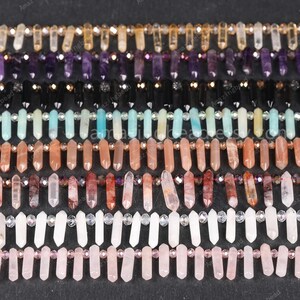 Può includere: Una collezione di fili di perline di pietre preziose in vari colori, tra cui viola, nero, turchese, pesca e rosa. Ogni filo presenta perline di cristallo allungate intervallate da perline più piccole e rotonde. Le perline sono disposte in file parallele su uno sfondo scuro.