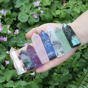 May include: A collection of eight crystal points in various colors, including clear, pink, purple, green, blue, and black. The points are arranged in a row on a hand, with the tips pointing towards the camera.