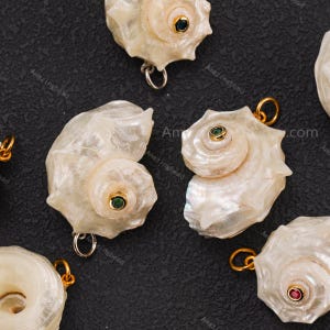 May include: A collection of iridescent white seashell pendants, each featuring a central gold-toned setting with a small embedded gemstone in colours including blue, green, and red, and a gold-toned metal jump ring attached to the top.