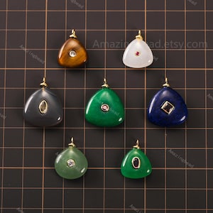 May include: A selection of gemstone pendants in shades of green, blue, and grey. Each pendant has a gold-coloured bail and features a central gemstone or crystal. The pendants are displayed on a grid background.