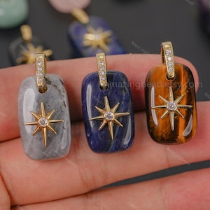 Pode incluir: Três pingentes retangulares de pedra nas cores cinza, azul profundo e olho de tigre marrom. Cada pingente apresenta um emblema de estrela em metal dourado com uma pedra central clara e um passador dourado com pequenos cristais.