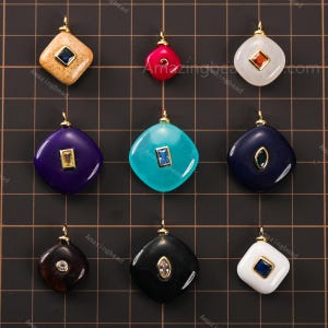 May include: A collection of nine gemstone pendants in various colors, including tan, red, white, purple, turquoise, and black. Each pendant has a gold-colored bail and a gemstone accent. The pendants are square with rounded corners.