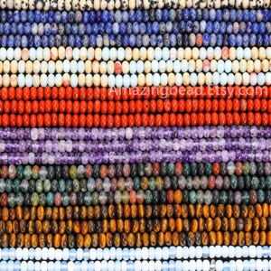 May include: A close-up of rows of gemstone beads in a variety of colours, including blue, white, red, purple, green, and brown. The beads are strung together, creating a textured pattern. The image features a variety of bead types and colours.