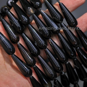 May include: A close-up shot of a string of black, teardrop-shaped beads. The beads are strung together, and the surface reflects light. The text "Amazingbead etsy.com" is visible.