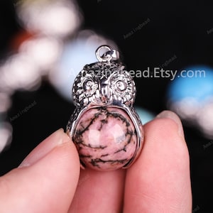 May include: A silver owl pendant featuring a pink and black rhodochrosite gemstone. The owl is holding the gemstone in its talons.