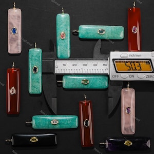 May include: Various rectangular gemstone pendants in shades of pink, green, red, and black. Each pendant has a small, oval gemstone and a gold-coloured bail. The digital caliper displays a measurement of 50.3 mm, equivalent to approximately 1.98 inches.