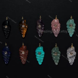 May include: A collection of gemstone pendants in the shape of seashells. Each pendant features a different color and stone type, including black, tiger's eye, amethyst, rose quartz, and turquoise. Each pendant has a small, round, clear gem in the center and a gold-colored bail.