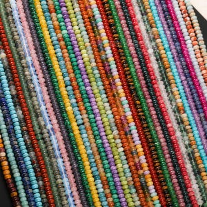 May include: A close-up view of numerous strands of colorful gemstone beads. The beads are small and round, in a variety of colors including blue, green, red, yellow, and purple. The beads are strung together, creating a textured and vibrant display.