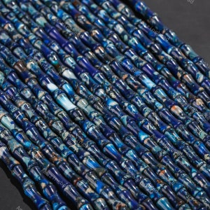 Könnte beinhalten: Eine Strähne aus blauen und weißen, gemusterten, zylindrischen Perlen. Die Perlen sind aus Naturstein und haben eine glatte, polierte Oberfläche.