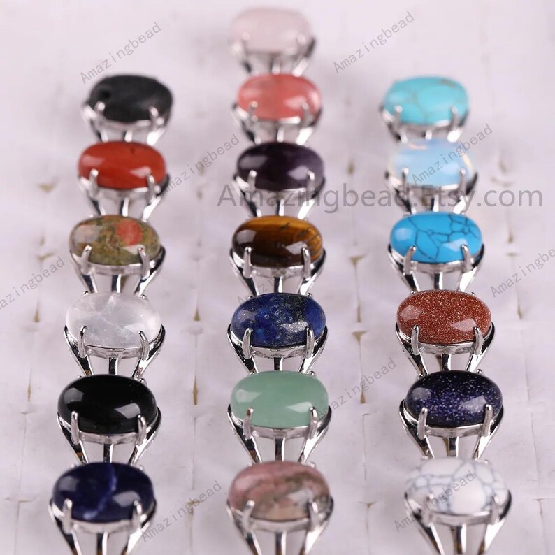May include: A set of 14 oval-shaped gemstone rings with silver settings. The gemstones are various colors, including red, green, blue, black, white, and brown. The rings are arranged in two rows, with seven rings in each row.