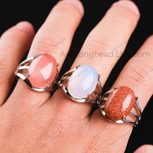 May include: Three silver rings with oval-shaped gemstones. The rings feature a delicate, open design. The gemstones are pink, white, and brown.