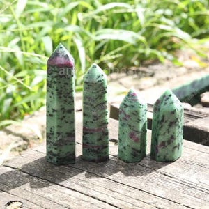Op de afbeelding: Vier groene en rode zoisiet kristallen obelisken van verschillende hoogtes zijn gerangschikt op een houten oppervlak. De kristallen hebben een gespikkelde uitstraling met donkergroene en rode insluitsels. De achtergrond heeft groen gebladerte.