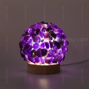 K&ouml;nnte beinhalten: Eine violette Amethyst-Kristalllampe mit einem Holzsockel. Die Lampe ist eingeschaltet und die Kristalle leuchten.