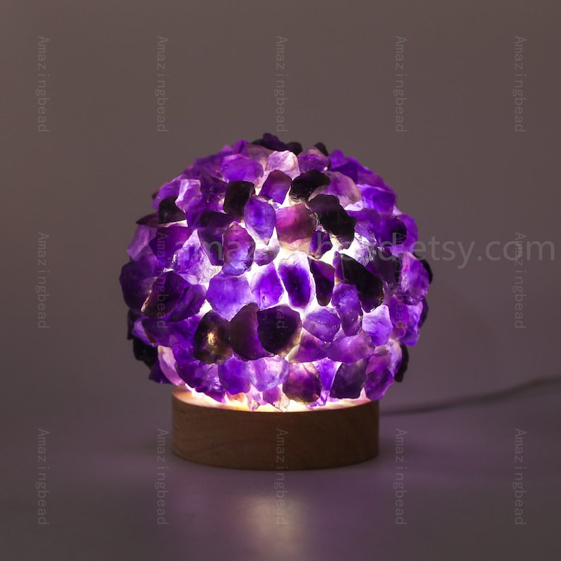 K&ouml;nnte beinhalten: Eine violette Amethyst-Kristalllampe mit einem Holzsockel. Die Lampe ist eingeschaltet und die Kristalle leuchten.