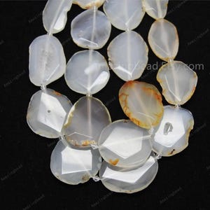 Puede incluir: Una hebra de cuentas de piedra de ágata blanca con forma ovalada irregular facetada. Las cuentas tienen un color blanco translúcido con inclusiones marrones y naranjas.