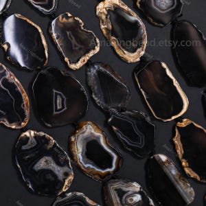 May include: A selection of polished black agate pendants, each with distinctive patterns and natural edges. The pendants display a range of shapes and sizes, featuring white, brown, and grey banding. The Etsy shop name "Amazingbead.etsy.com" is visible on some pendants.