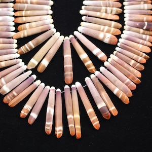 Puede incluir: Un collar compuesto por numerosos elementos cilíndricos alargados. Los elementos son principalmente en tonos morados, beige y naranja, con rayas blancas. El collar está dispuesto en forma de arco sobre un fondo negro.