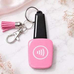May include: A pink contactless payment keychain shaped like a nail polish bottle with a black cap. It features a white circular icon with signal waves in the center. The keychain includes a silver metal ring, a silver lobster clasp, and a pink tassel.