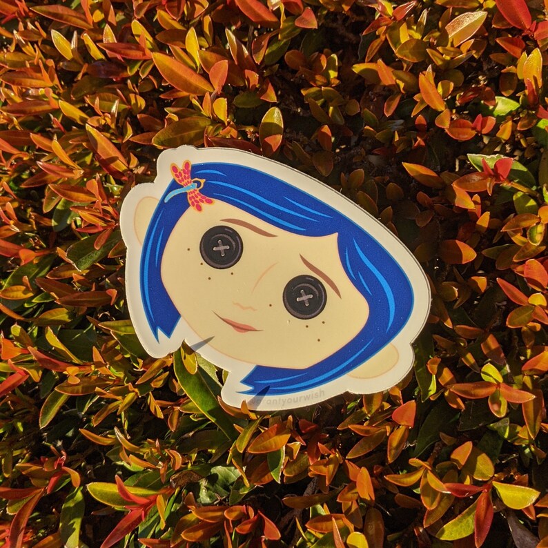 May include: A sticker featuring a cartoon character with blue hair and large, button-like eyes. The character has a red and white butterfly on her hair. The sticker is on a green leafy background.