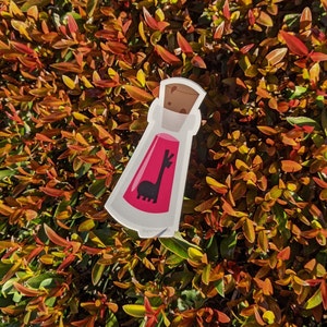 May include: A pink potion bottle sticker with a black llama silhouette inside. The sticker is on a background of red and green foliage.