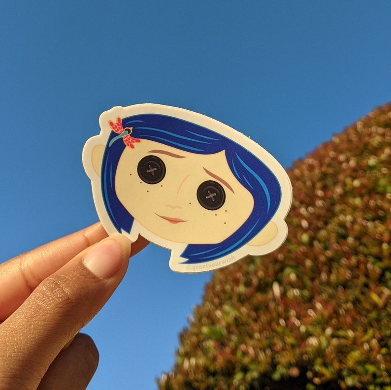 May include: A sticker featuring a cartoon illustration of a girl with blue hair and large, button-like eyes. She is wearing a red dragonfly in her hair. The sticker has a white background and the text "@grantyourwish" is printed at the bottom.