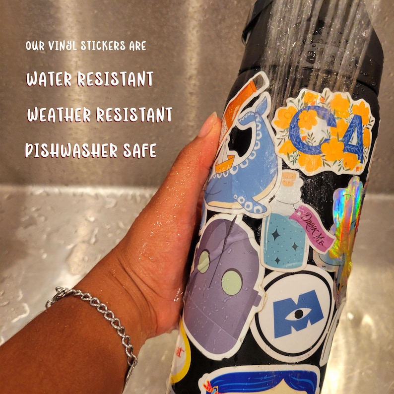 May include: A black reusable water bottle covered in colorful vinyl stickers. The stickers feature a whale, a robot, a monster eye, a floral design, and a cactus. The text "Drink Me" is visible on one of the stickers. The bottle is being rinsed under running water in a stainless steel sink. The text "OUR VINYL STICKERS ARE WATER RESISTANT WEATHER RESISTANT DISHWASHER SAFE" is visible above the bottle.