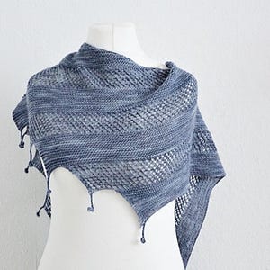 Peut inclure: Un châle tricoté triangulaire dans des tons variés de bleu et de gris. Le vêtement présente des sections alternées de point mousse et de motifs ajourés, terminées par de petites houppes décoratives le long du bord inférieur du tissu.