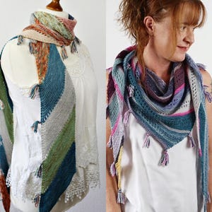 Pode incluir: Um xale de tricô triangular com listras diagonais em tons de azul, verde, cinza e rosa. O acessório é feito com um padrão de ponto texturizado e finalizado com pequenas borlas de fio multicoloridas ao longo das bordas externas.
