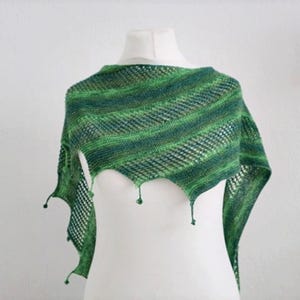 Könnte beinhalten: Ein grün-blauer Strickschal mit diagonalem Streifenmuster. Der Schal ist über einer weißen Schaufensterpuppe drapiert. Die Kanten des Schals haben ein gewelltes Design mit kleinen grünen Quasten.