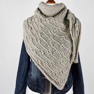 May include: A triangular knit shawl in a light beige colour featuring a textured pattern of diagonal lines and small raised knots. The shawl is draped over a dark blue denim jacket, showcasing the intricate stitch work and the soft yarn texture.