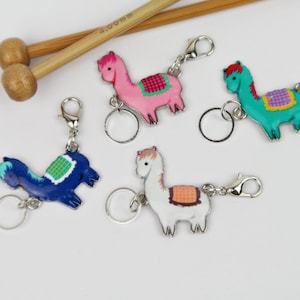 Könnte beinhalten: Vier farbenfrohe Lama-förmige Maschenmarkierer mit silbernen Schlüsselringen und Karabinerhaken. Die Maschenmarkierer sind rosa, blau, grün und weiß mit gestickten Details.