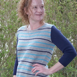 Peut inclure: Un pull gris avec des rayures horizontales bleues et bleu clair. Les manches sont bleu marine uni. Le pull est porté par une personne.