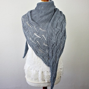 Könnte beinhalten: Ein dreieckiges Stricktuch in staubigem Blau mit einer Kombination aus kraus rechts gestrickter Textur und komplizierten Spitzenmustern mit Lochmusterdetails. Das Tuch liegt über einem weißen Oberteil mit einem Saum aus Spitze.