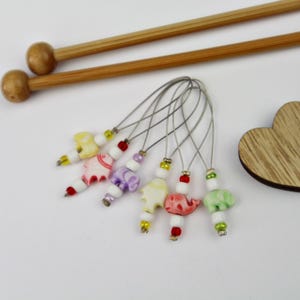 Könnte beinhalten: Eine Sammlung farbenfroher, elefantenförmiger Maschenmarkierer mit Stricknadeln aus Holz. Die Maschenmarkierer sind in verschiedenen Farben gehalten, darunter Gelb, Rosa, Lila und Grün, mit weißen und roten Perlen. Die Stricknadeln sind aus hellbraunem Holz.
