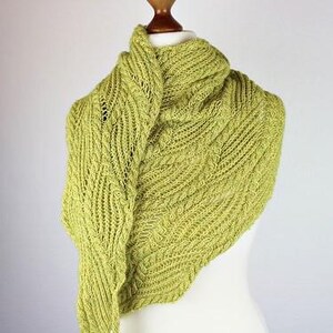 Könnte beinhalten: Ein hellgrüner Strickschal, der auf einer Schaufensterpuppe präsentiert wird. Der Schal hat ein strukturiertes, gemustertes Design und eine dreieckige Form. Das Garn wirkt weich und der Gesamtstil ist gemütlich und handgefertigt.